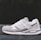 A white and gray athletic shoe with a modern design is positioned on a flat surface against a blurred dark background. The shoe features a mix of materials, including mesh and suede, and showcases detailed stitching with a visible logo and embossed text on the side.