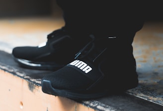 A close-up view of black athletic shoes with a visible brand logo, placed on a concrete step. The shoes are stylishly designed and appear to be suitable for sports or casual wear. The background is blurred, focusing attention on the footwear.