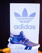 A sneaker displayed in front of a branded backdrop. The shoe has a modern design with a combination of blue, white, and green accents. The background features a large logo and contrasting colors, adding to the focus on the sneaker.