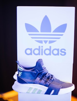 A sneaker displayed in front of a branded backdrop. The shoe has a modern design with a combination of blue, white, and green accents. The background features a large logo and contrasting colors, adding to the focus on the sneaker.