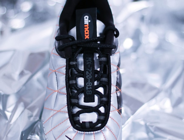 A single sneaker with a sleek, modern design lies on a metallic, crumpled background. The shoe features a white base with black laces and accents, as well as orange stitching detail. The brand's name is visible on the tongue of the shoe.