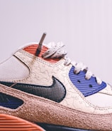 A close-up view of a stylish sneaker featuring a prominent swoosh logo on the side. The design includes textured patterns on a beige upper with navy and red details. The sole has a brown, rugged appearance.