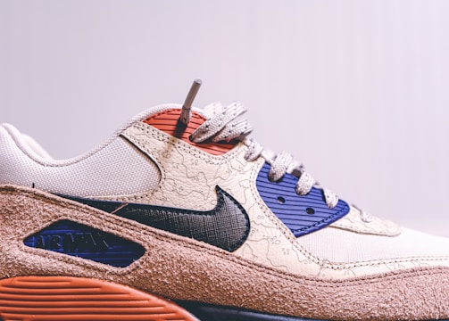 A close-up view of a stylish sneaker featuring a prominent swoosh logo on the side. The design includes textured patterns on a beige upper with navy and red details. The sole has a brown, rugged appearance.