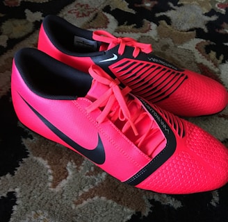 A pair of bright red athletic shoes with black accents and laces are placed on a patterned carpet. The shoes have a sleek, modern design with textured detailing and branding visible on the sides.