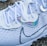 A close-up of a stylish white athletic shoe with intricate patterns and shoelaces. The shoe features a sleek design with iridescent accents near the eyelets and on a prominent logo.