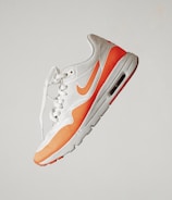 A stylish athletic sneaker featuring a white and orange color scheme, with perforated detailing and a visible air cushion in the sole. The shoe is positioned against a plain background, emphasizing its sleek and modern design.