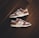 A pair of brown and white sneakers with prominent branding positioned on a dark wooden floor. The stylish shoes have a low-top design with white shoelaces and a distinctive swoosh logo on the sides.