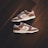 A pair of brown and white sneakers with prominent branding positioned on a dark wooden floor. The stylish shoes have a low-top design with white shoelaces and a distinctive swoosh logo on the sides.