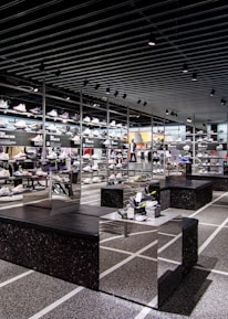 A modern sneaker store with sleek black shelving displaying various athletic shoes and sports apparel. Central seating areas and spotlights provide a contemporary retail ambiance. The signage prominently features a famous sportswear slogan in the background.