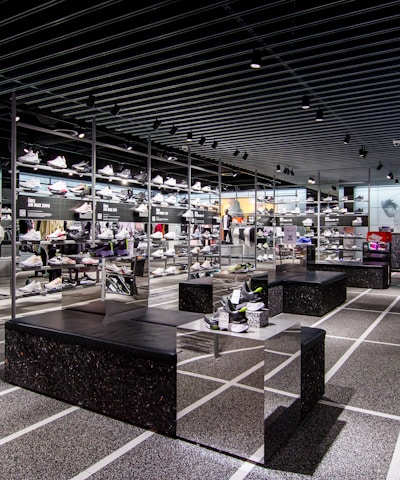 A modern sneaker store with sleek black shelving displaying various athletic shoes and sports apparel. Central seating areas and spotlights provide a contemporary retail ambiance. The signage prominently features a famous sportswear slogan in the background.