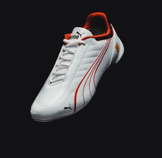 A sleek white sports shoe with red accents and logo details, featuring a textured surface and clean design lines. The background is dark, emphasizing the shoe.