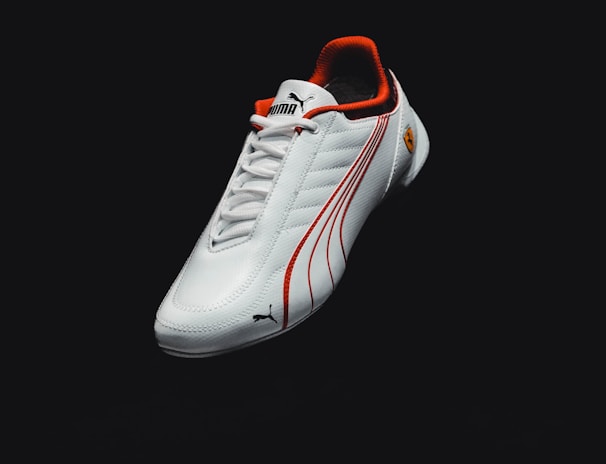 A sleek white sports shoe with red accents and logo details, featuring a textured surface and clean design lines. The background is dark, emphasizing the shoe.