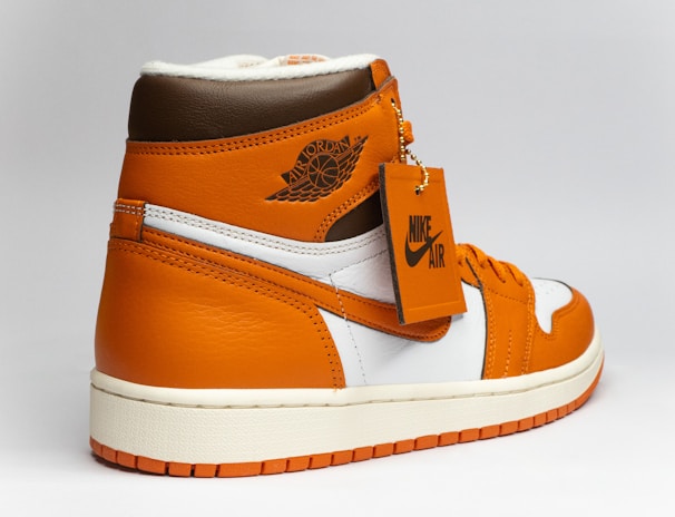 A stylish athletic shoe with a high-top design, featuring a combination of orange, brown, and white colors. The shoe displays a prominent logo on the side and a tag attached to the laces.