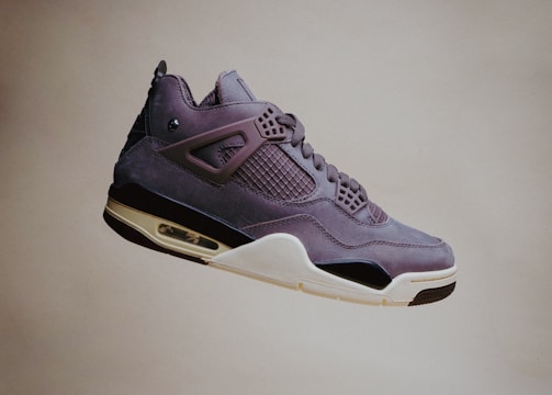 A single sneaker with a sleek, modern design is visibly suspended in mid-air against a plain background. The shoe features a mix of materials, including smooth leather and mesh, with intricate detailing around the lace area and a distinctive air cushion in the sole.