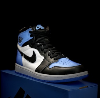 A stylish athletic shoe is placed on a shoebox, featuring a combination of black, white, and light blue. The shoe design includes a prominent logo on the side and a high-top silhouette.