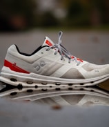 A white and red athletic shoe is positioned on a reflective surface, showing a clear reflection of the shoe. The shoe has a unique textured design with visible branding and design elements on its side. The background is slightly blurred, giving emphasis to the shoe and its reflection.